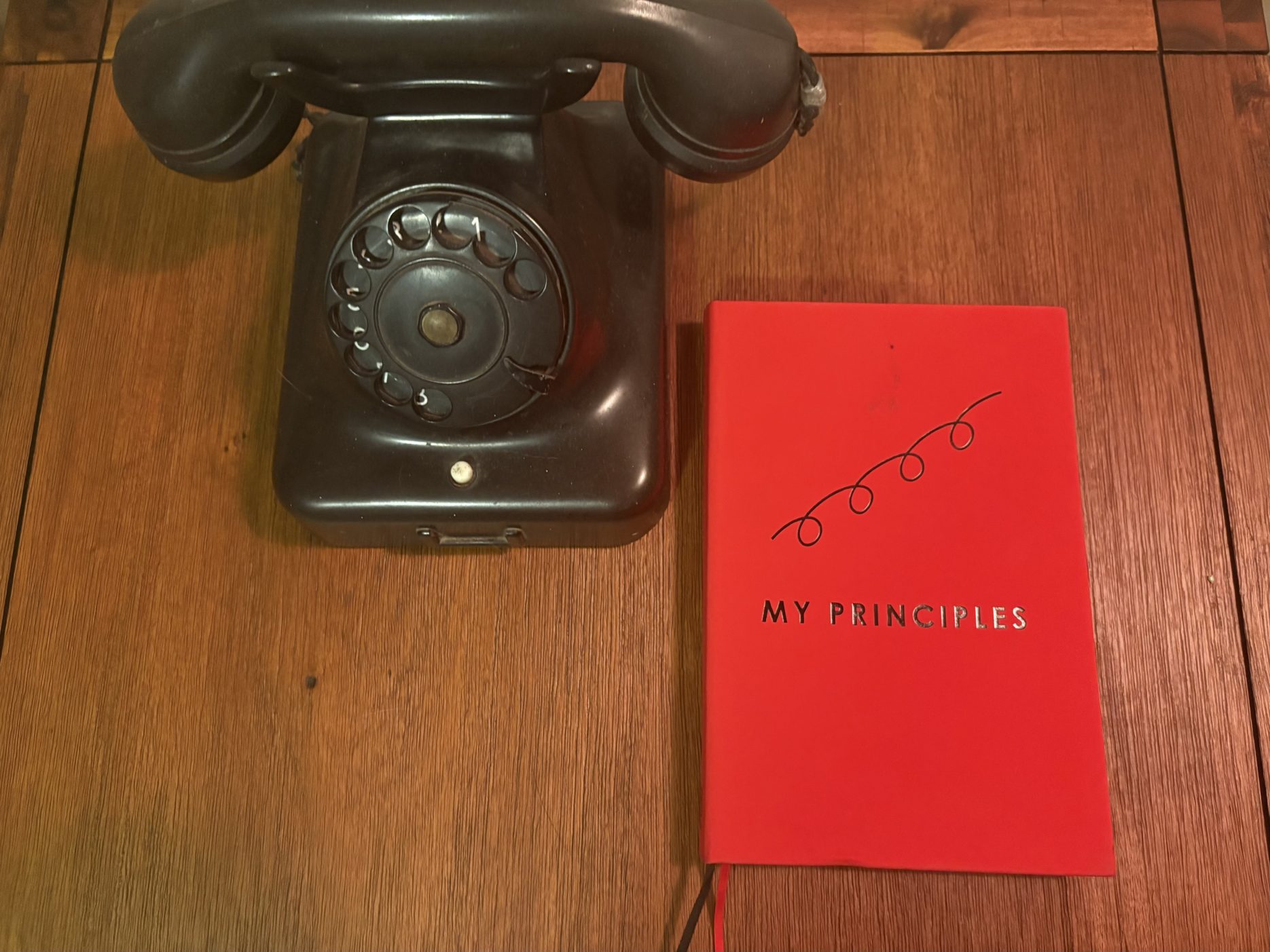 An overhead shot shows a vintage black rotary telephone and a vibrant red journal resting on a polished wooden table. The phone, positioned on the left, features a classic curved handset and a circular dial with white numbers. To its right lies the journal, which has a minimalist cover featuring a simple black curly line illustration above the words "MY PRINCIPLES" printed in a clean, sans-serif font. The warm lighting highlights the textured grain of the dark wood surface beneath them.