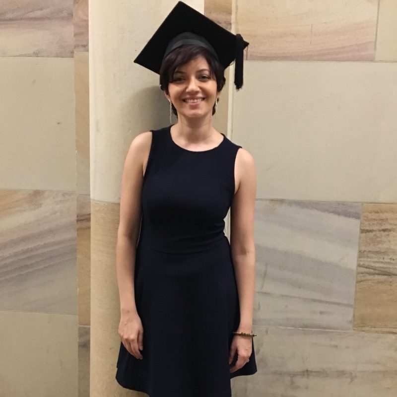 A smiling woman in a navi blue dress and academic graduation cap, standing against the iconic light-coloured sandstone walls of the University of Queensland (UQ) during her graduation ceremony eight years ago.