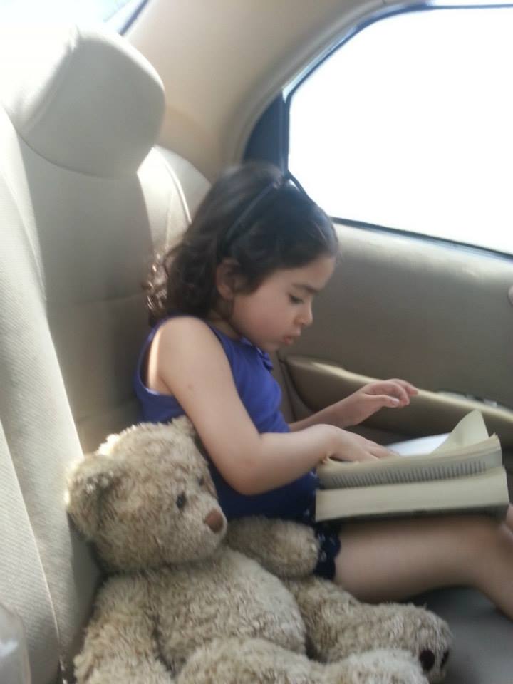 Syma Ahmed's daughter reading a book in a car with her teddy bear by her side. 