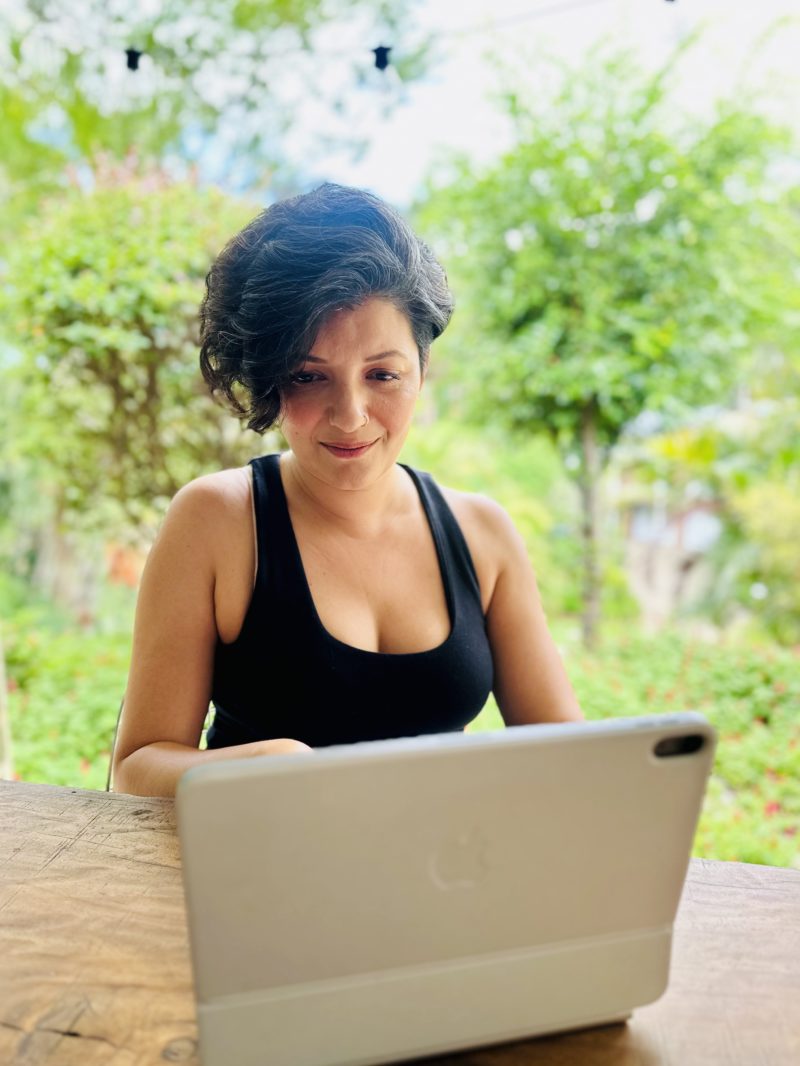 Portrait of Syma Ahmed in a moment of deep focus, working on her laptop in a garden setting.