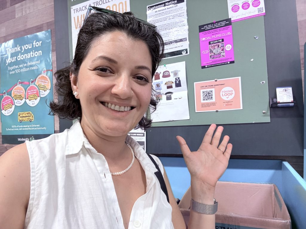 A close-up selfie of Syma Ahmed standing in front of a community board in Brisbane after pinning a Found Loop poster on a community notice board. 